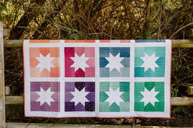 Joyful Stars quilt in vibrant ombre colors, draped over a wooden fence. The quilt features a modern star block design in shades of orange, pink, purple, blue, and green, with a white background and straight-line quilting.