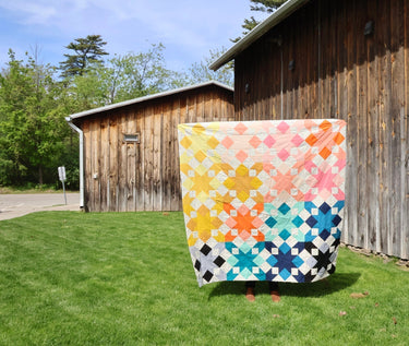 Completed Star Struck star quilt top in a rainbow colorway held up on a sunny lawn showing the full quilt size against a rustic barn