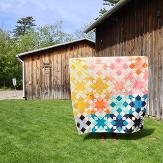Completed Star Struck star quilt top in a rainbow colorway held up on a sunny lawn showing the full quilt size against a rustic barn