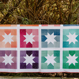 Joyful Stars quilt in vibrant ombre colors, draped over a wooden fence. The quilt features a modern star block design in shades of orange, pink, purple, blue, and green, with a white background and straight-line quilting.