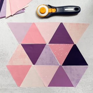 Pink and purple 60° triangle quilt pieces arranged with a rotary cutter, part of Cotton and Joy's modern quilting tutorial for beginners.