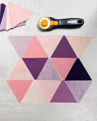 Pink and purple 60° triangle quilt pieces arranged with a rotary cutter, part of Cotton and Joy's modern quilting tutorial for beginners.