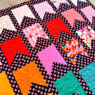 Detailed view of the Bookmarks quilt by Cotton and Joy, using Ruby Star Society fabrics in pink, red, orange, yellow, and teal bookmark-shaped blocks, arranged in a rainbow layout against a black background with pink daisy prints.