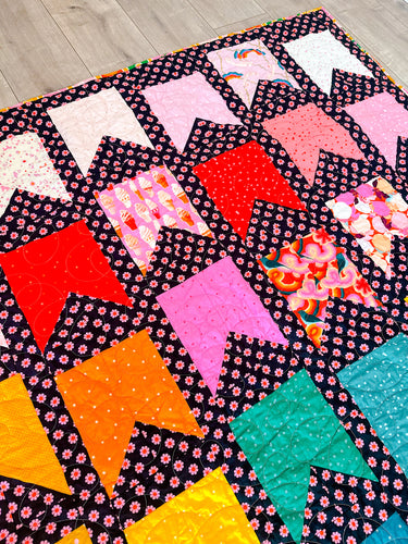 Detailed view of the Bookmarks quilt by Cotton and Joy, using Ruby Star Society fabrics in pink, red, orange, yellow, and teal bookmark-shaped blocks, arranged in a rainbow layout against a black background with pink daisy prints.