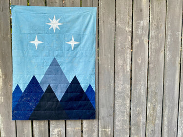 Corner of the Night Court quilt pattern by Cotton + Joy, showcasing a blue and black mountain design with metallic speckles, displayed against a wooden lattice background.