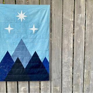 Corner of the Night Court quilt pattern by Cotton + Joy, showcasing a blue and black mountain design with metallic speckles, displayed against a wooden lattice background.