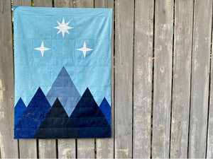 Corner of the Night Court quilt pattern by Cotton + Joy, showcasing a blue and black mountain design with metallic speckles, displayed against a wooden lattice background.